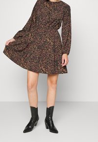 Robe noire à manches longues avec un motif abstrait coloré, taille cintrée et jupe évasée. Modèle portant des bottines noires brillantes à bouts pointus.