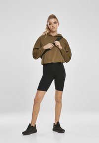 Sudadera corta de color verde oliva con mangas largas, combinada con pantalones cortos de ciclismo negros. Modelo lleva zapatillas deportivas negras y sostiene una correa negra.