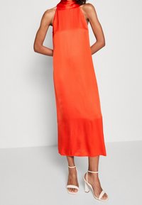 Frau in einem ärmellosen, leuchtend orangefarbenen Neckholder-Midi-Kleid mit weißen Knöchelriemen und Blockabsätzen, die vor einem einfarbigen hellen Hintergrund steht.