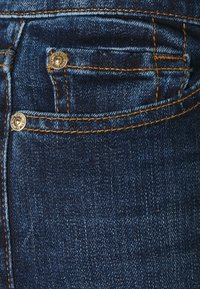 Jeans in denim blu scuro con tasca anteriore, cuciture giallo oro e tessuto testurizzato. Due rivetti metallici adornano l'area della tasca.