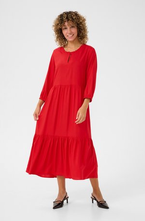 Robe midi rouge avec un col rond, des détails froncés, des manches longues et une jupe à volants. Associée à des chaussures à talons noires. Tissu lisse.