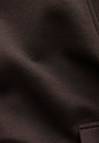 Tissu brun foncé à la texture lisse, présentant un motif tissé subtil. Une petite poche avec des détails de couture est visible sur le bord inférieur.