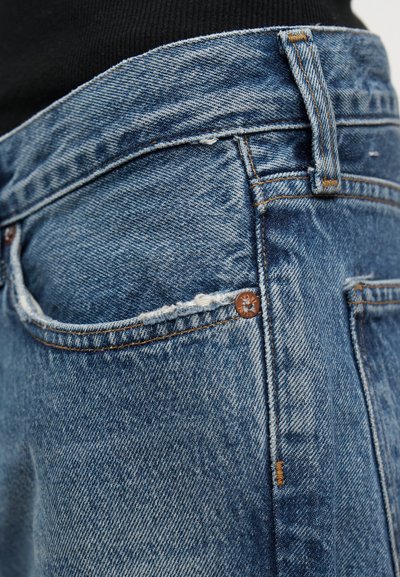 Κοντινή λήψη μπλε τζιν με εστίαση στην μπροστινή τσέπη, τη θηλιά για τη ζώνη και τις λεπτομέρειες από χάλκινο καρφί με φόντο μαύρο στο πάνω μέρος.