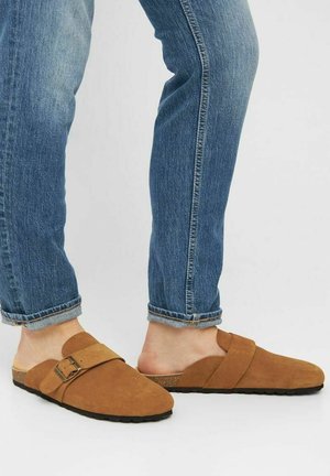 Brune ruskindssko i slip-on-stil med dobbelt rem og justerbar spænde, parret med opfoldede blå denimjeans, som viser fødderne.