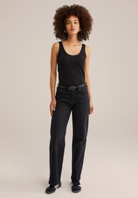 Nainen, jolla on kiharat hiukset, pukeutuneena mustaan toppiin, mustiin korkeavyötäröisiin farkkuihin, mustaan vyöhön ja mustiin lenkkareihin, seisoo neutraalia taustaa vasten.