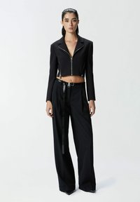 Crna skraćena jakna sa patentnim zatvaračem sprijeda, u kombinaciji s crnim hlačama visokog struka koje imaju sjajni pojas i detalj vertikalnih pruga.