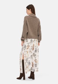 Brauner gestrickter Pullover mit hohem Kragen, kombiniert mit einem langen cremefarbenen Rock mit Tierillustrationen und seitlichen Schlitzen, getragen mit braunen Stiefeln.