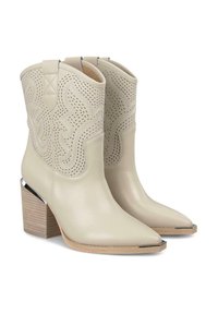 Beżowe skórzane botki za kostkę z szpiczastymi noskami, dekoracyjnymi wycięciami i masywnym drewnianym obcasem. Srebrne akcenty na nosku i obcasie podkreślają detale.