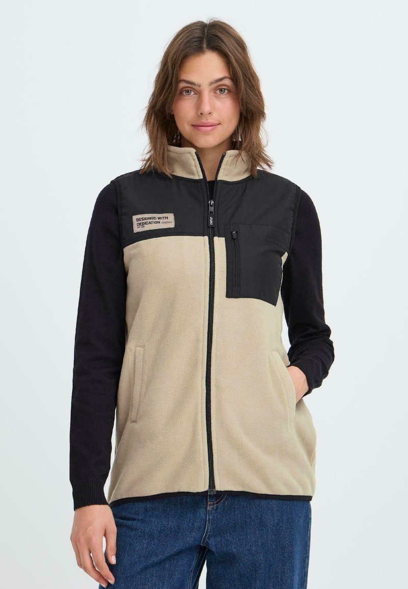Fleece-Weste in Beige und Schwarz. Verfügt über einen Reißverschluss vorne, seitliche Taschen und eine Brusttasche. Der Stoff hat eine weiche Textur mit einem kontrastierenden Design.