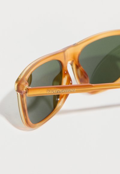 Διαφανή κεχριμπαρένια γυαλιά ηλίου με σκούρους πράσινους φακούς, που φέρουν το λογότυπο Dolce & Gabbana στο βραχίονα του σκελετού.