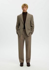 Brun rutete blazer med hakket slag, kombinert med en mørk poloskjorte og lysebrune bukser. Glatt tekstur, skreddersydd passform og subtil glans.