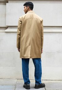 Khaki trench coat dengan potongan lurus, dilengkapi dengan lengan panjang dan ventilasi di bagian belakang, dipadukan dengan celana jeans denim biru dan sepatu hitam.