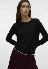 Svart cardigan med långa ärmar, rund halsringning och knäppning framtill, med en kontrasterande off-white kant vid nederkanten.