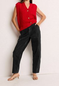 Punainen neulepaita ilman hihoja, jossa on timanttikuvio ja puunapit, yhdistettynä löysiin mustiin farkkuihin ja vaaleanruskeisiin korkokenkiin.