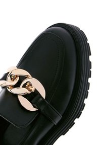 Zwarte leren schoen met een stevige zool, voorzien van een glanzende gouden kettingaccent en brogue-details op de bovenkant. Zachte textuur, modern ontwerp.