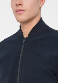 Námořnická bombardér bunda s žebrovaným límcem, hladkou texturou a předním zipem. Má minimalistický design, bez výrazných vzorů nebo akcentů.