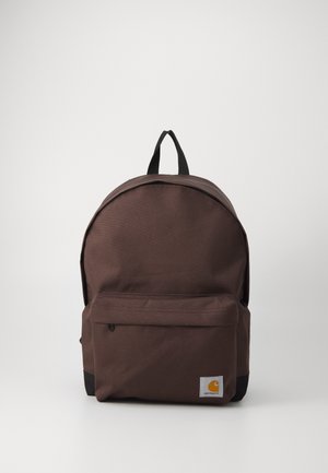 Mochila de lona marrón con una parte superior redondeada, bolsillo frontal con cremallera, acento negro en la parte inferior y un parche de logo prominente en el frente.
