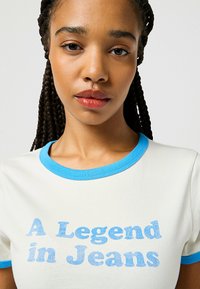 Camiseta blanca de manga corta con cuello acanalado en azul claro y texto impreso "Una Leyenda en Jeans" en una suave y texturizada fuente azul claro.