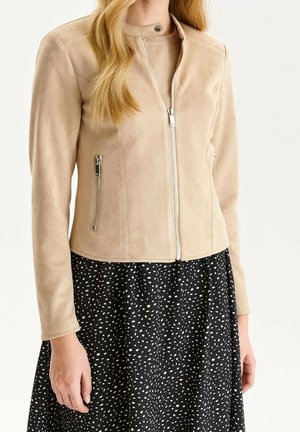 Woman wearing a beige zip-up suede jacket over a black skirt with white irregular dot pattern, standing against a plain background.
