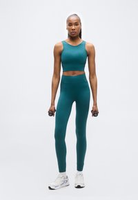 Γυναίκα με αθλητική στολή σε απόχρωση του τιρκουάζ, όρθια, κρατώντας αλτήρες 1 κιλού σε κάθε χέρι, με φόντο απλό λευκό.