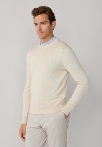 Pull beige clair à col rond, confectionné dans un tissu doux. Comprend des poignets et un ourlet côtelés, associé à une chemise bleu clair à col en-dessous.
