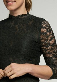 Schwarzes Spitzentop mit hochgeschlossenem Kragen und drei Viertel langen Ärmeln, das mit floralen Mustern und einem gewellten Saum verziert ist.