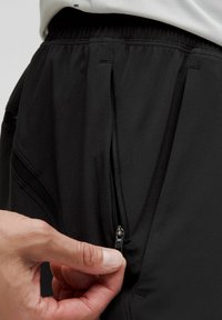 Hand som håller i dragkedjan på fickan till svarta träningsbyxor med elastisk midja och nätdetaljer på sidan.