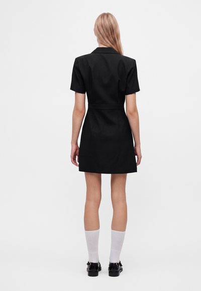 Femme debout de dos, vêtue d'une robe noire courte à manches courtes, de chaussettes blanches et de chaussures noires sur un fond uni.