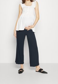 Bílý bezramenný top s volánkovými ramínky a detailem knoflíků, kombinovaný s volnými, tmavě modrými širokými kalhotami a černými nazouvacími botami.