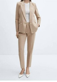 Costume beige sur mesure avec un blazer croisé, des revers en pointe et deux boutons. Un pantalon ajusté complète l'ensemble, associé à des chaussures blanches à pointes.