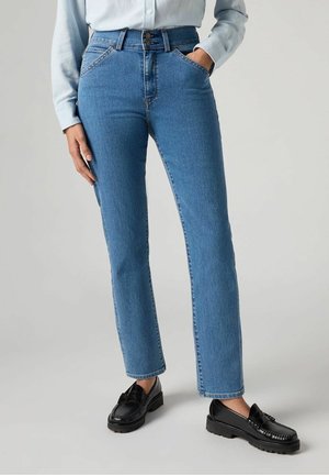 Osoba ubrana w jasnobłękitną koszulę zapinaną na guziki, niebieskie proste dżinsy z wysokim stanem i czarne mokasyny, stojąca z jedną ręką w kieszeni.