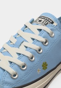 Nærbillede af lyseblå Converse-sneaker med hvide snørebånd og broderede grønne og hvide blomster nær sålen.