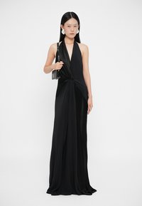 Čierne šaty s halter krkom a prekrúteným predným dizajnom, vyrobené z hladkej látky. Modelka drží malú čiernu kabelku a stojí na bielom pozadí.