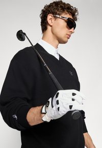 Čierny sveter s V-výstrihom, biela košeľa s golierom pod ním, drží golfovú palicu. Biele golfové rukavice s čiernymi akcentmi, nosí slnečné okuliare.