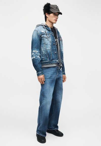 Νεαρός άνδρας όρθιος, φορώντας ένα φθαρμένο μπλε τζιν μπουφάν πάνω από γκρι φούτερ με κουκούλα, τζιν με φαρδιά πόδια, μαύρα παπούτσια και σκουρόγκρι καπέλο με καλύμματα για τα αυτιά.