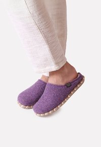 Pehmeät violetit tossut, joissa on pyöreä kärki, kudottu tekstuuri pohjassa ja pehmeä sisävuori. Logo-tunnus näkyvissä sivulla.