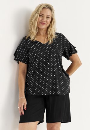 Lyshåret kvinde iført sorte shorts og sort polkaprikbluse med korte ærmer står mod en hvid væg og smiler med hånden på hoften.