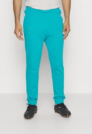 Homme portant un pantalon de survêtement turquoise avec le logo du FC Barcelone et des chaussures de football noires, debout devant un fond blanc uni.