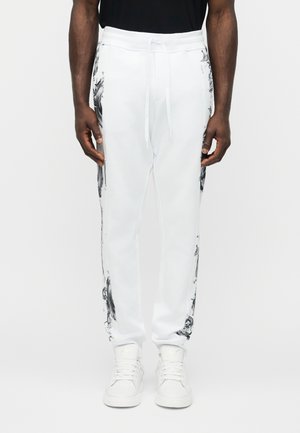 Άνδρας που φοράει λευκό φόρμα με μαύρα αφηρημένα σχέδια στο πλάι και λευκά αθλητικά, στέκεται μπροστά σε ένα απλό φόντο.