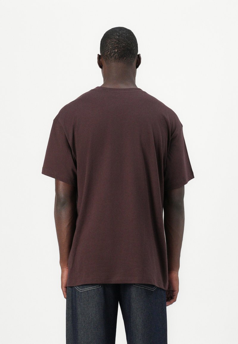 T-shirt in cotone marrone con maniche corte, vestibilità rilassata e colletto tondo. Presenta una texture liscia e nessun motivo o grafica visibile.