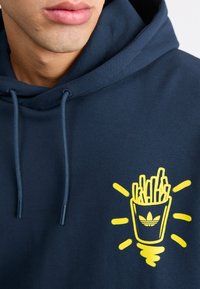 Laivaston huppari, joka on valmistettu puuvillasekoitteesta; siinä on keltainen graafinen kuva ranskalaisista perunoista kupissa Adidas-logolla, pyöreät vetoketjut ja ribbaatut hihansuut.