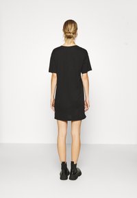 Une femme se tient de dos, portant une robe noire à manches courtes et des bottines noires, les cheveux attachés en un chignon bas.