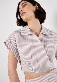 Camisa corta a rayas en beige claro y blanco. Consta de mangas cortas, un cuello y dos bolsillos frontales. Tela suave y texturizada.