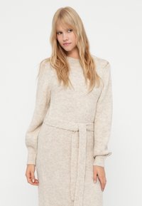 Abito lavorato a maglia color beige chiaro con vita a corsetto, scollatura tonda e lunghe maniche a sbuffo, che presenta un motivo morbido e testurizzato.