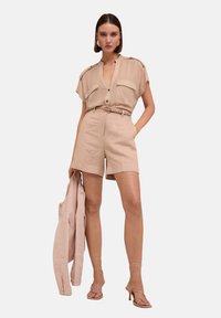Beige kortärmad skjorta med knäppning fram, två bröstfickor, matchande bälte och skräddarsydda beige shorts. Modellen håller en lätt jacka och har på sig sandaletter med remmar.
