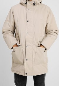 Parka acolchada beige con capucha desmontable, botones a presión delanteros y dos grandes bolsillos laterales. Textura suave y reguladores ajustables.