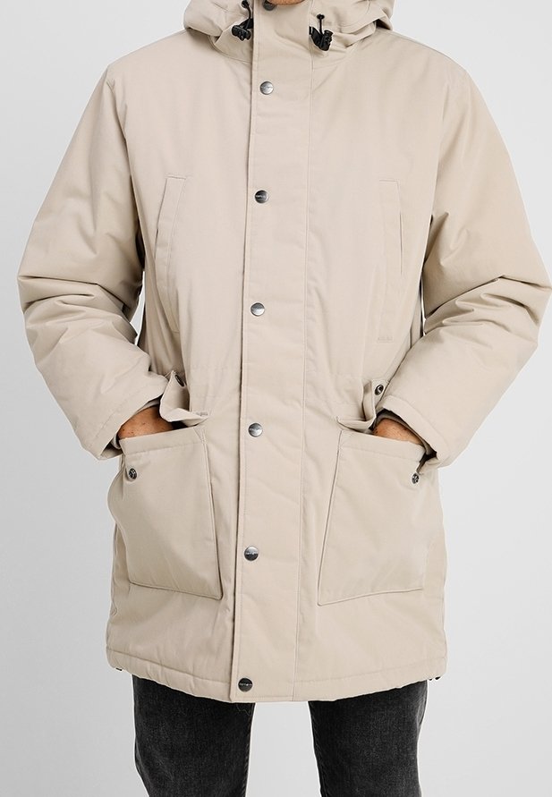 Parka acolchada beige con capucha desmontable, botones a presión delanteros y dos grandes bolsillos laterales. Textura suave y reguladores ajustables.