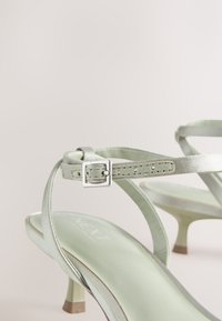 Sandales à talons hauts vert clair avec des fines brides et une petite boucle argentée sur un fond clair uni.