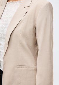 Ženska, ki nosi bež blazer preko bele čipkaste majice, prikazano so podrobne teksture in šivi na blazerju.