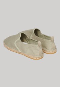 Zapatillas de lona tipo slip-on en verde oliva con suela de yute trenzada, que cuentan con paneles elásticos en los laterales y una etiqueta de tela en el talón.